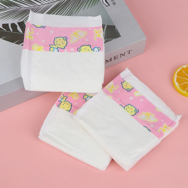 Três fraldinhas Reborn com banda rosa estampada, dispostas em leque sobre caixa cinzenta e fatia de laranja, fundo rosa.